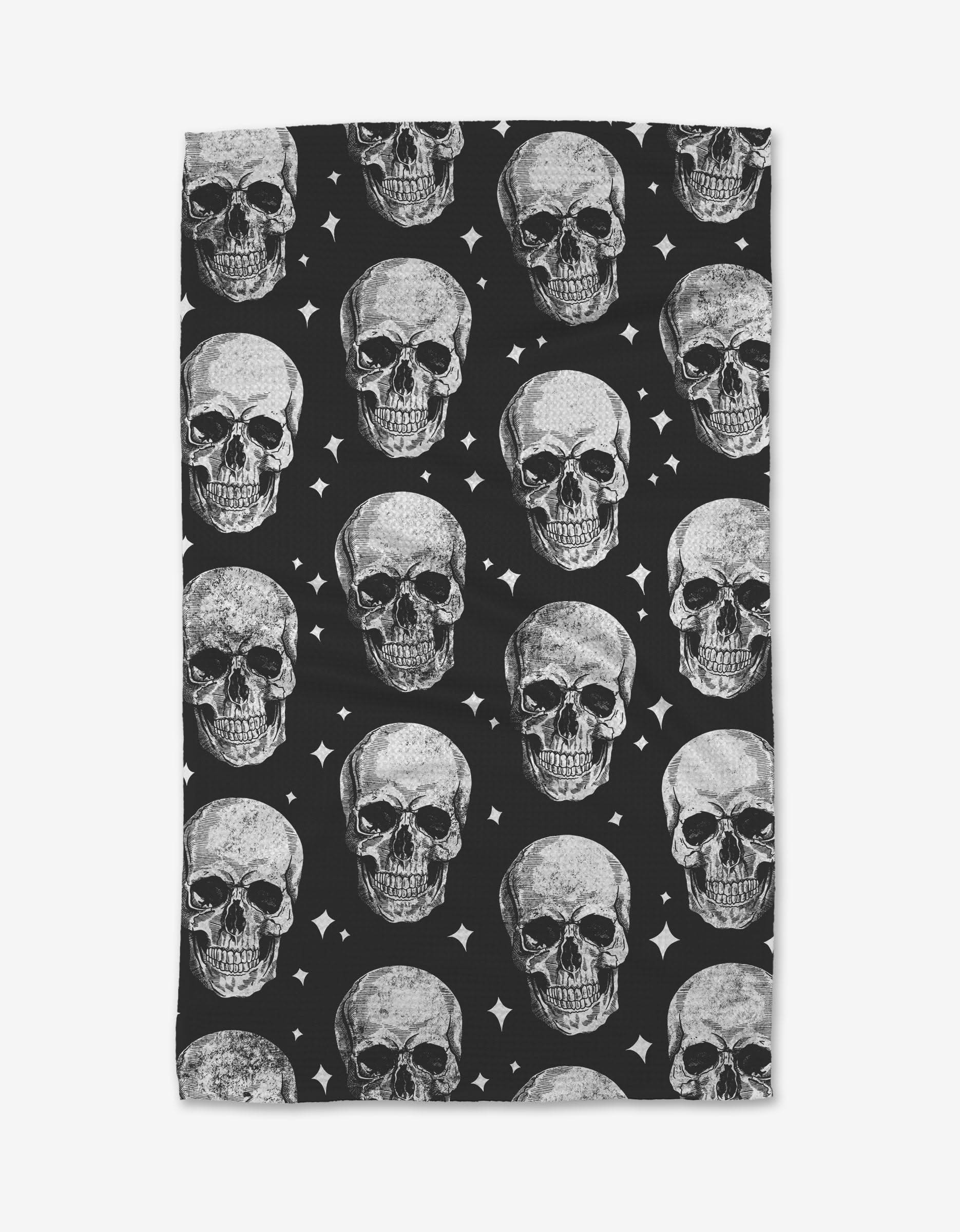 The GEOMETRY Skulls tea towel is rectangular, made from recycled materials, and features a repeating pattern of realistic gray skulls on black with small white star-like accents.