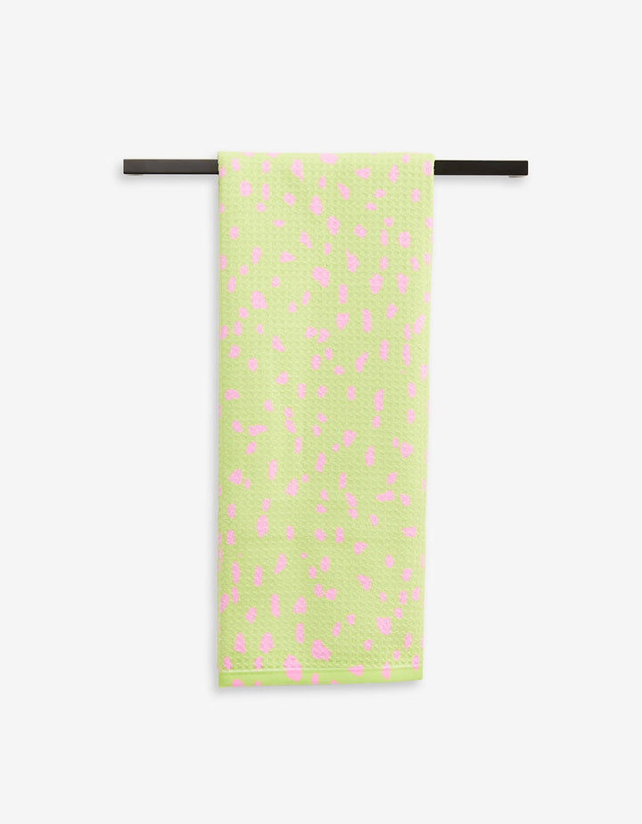 The GEOMETRY Speckle Green tea towel, featuring light green Geoweave fabric with pink spots, hangs neatly on a black towel bar against a white background.