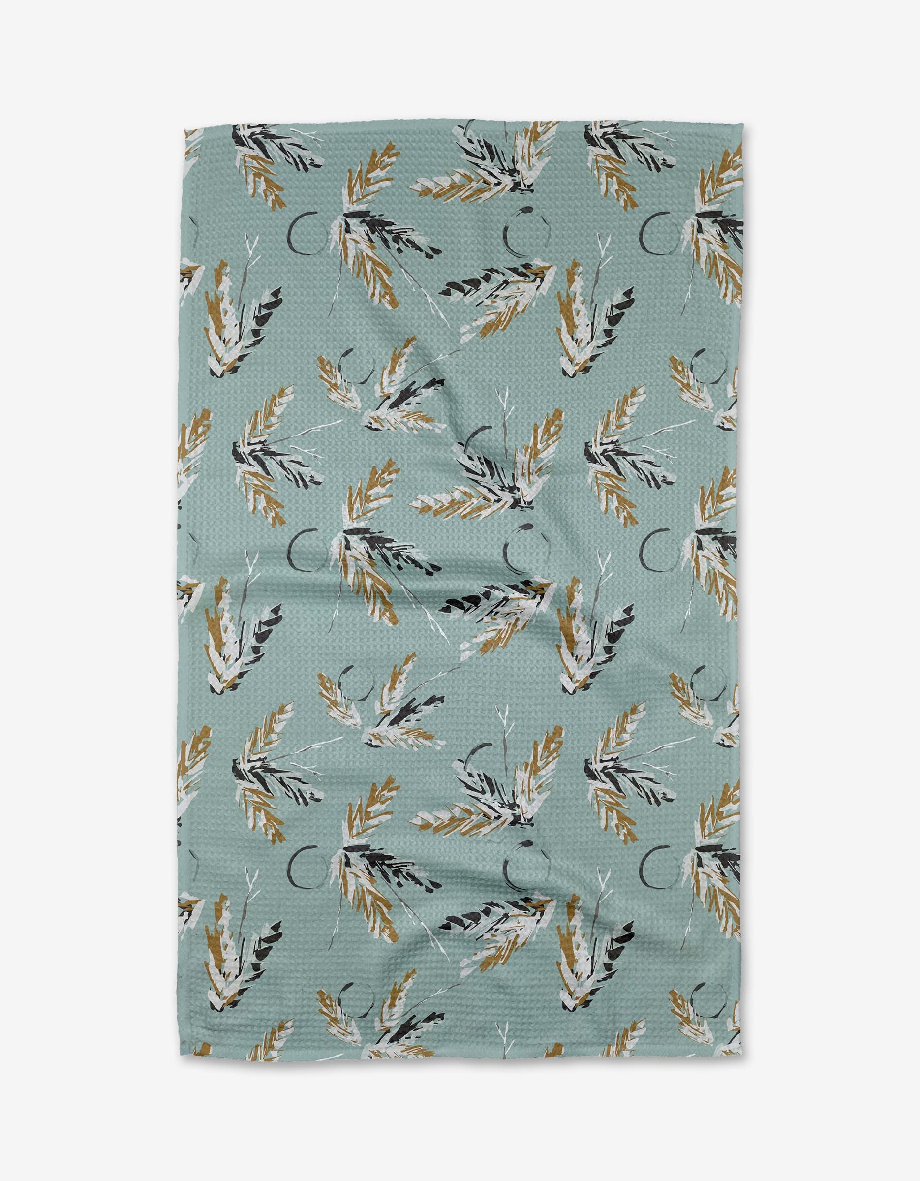 The Surround tea towel by Toboggan Avenue is rectangular, light blue-green, and made from quick-drying recycled materials. It features yellow, white, and black leaves scattered across its slightly textured fabric.