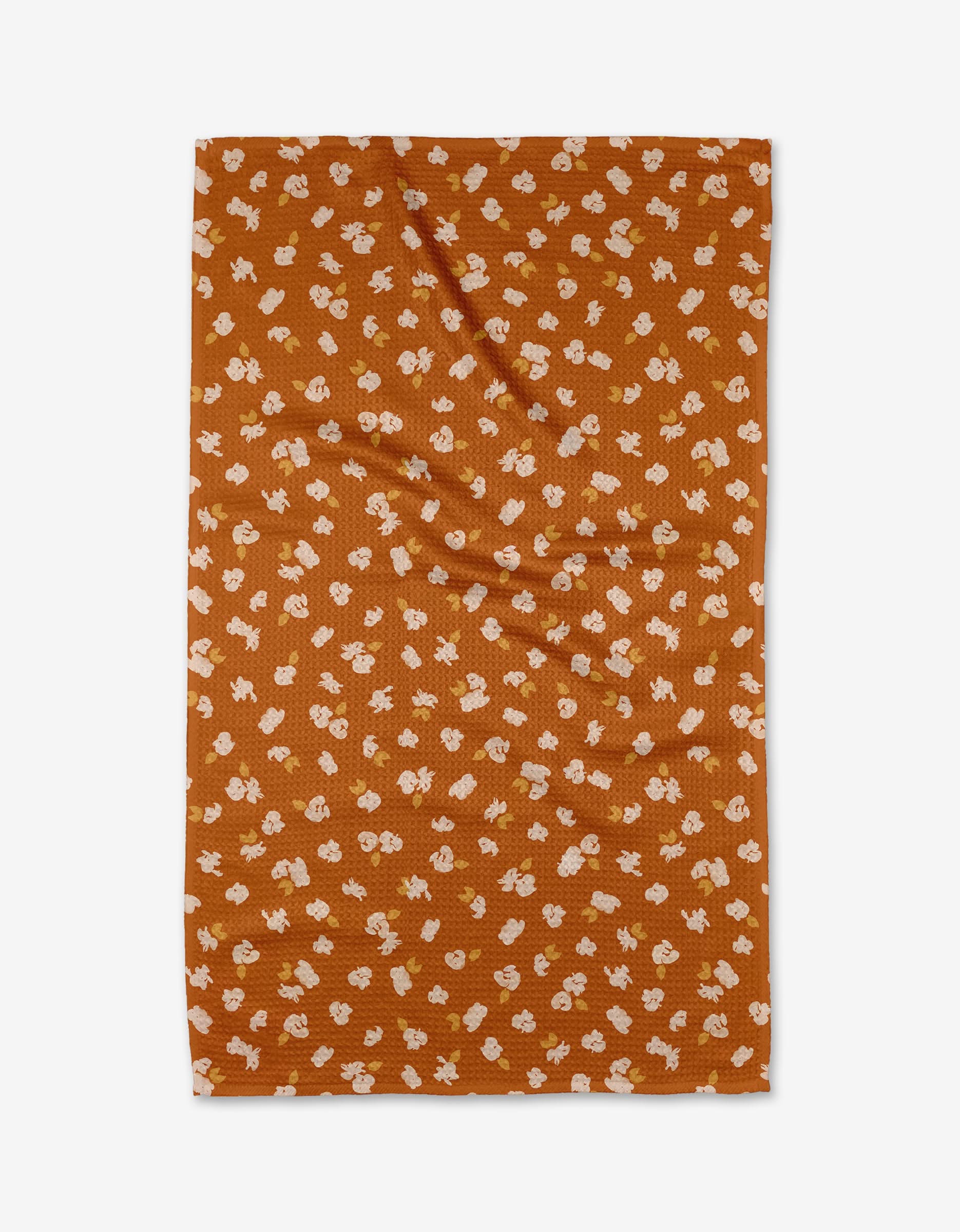 The Tossed Wildflowers Rust tea towel by Mandy Arroyo is rectangular, rust-colored, features small white and yellow flowers on Geoweave fabric, and is shown laid flat on a white background.