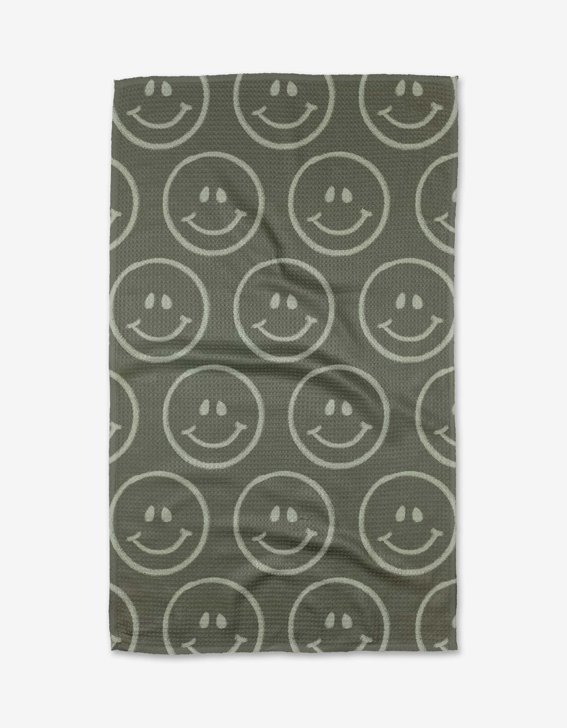 The You Make Me Happy tea towel by Kelsi Fullmer features a gray Geoweave fabric with large, white outlined smiley faces arranged in rows. The rectangular towel has a slightly wrinkled look.